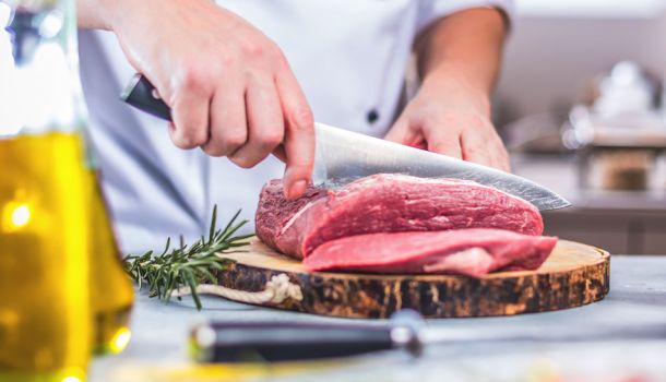 Person in weißer Kochjacke schneidet rohes Rindfleisch auf einem Holzbrett, daneben liegen Rosmarinzweige.