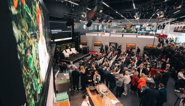 In einer Messehalle sitzen viele Menschen auf Stühlen vor einer Bühne, auf der leere Stühle stehen.