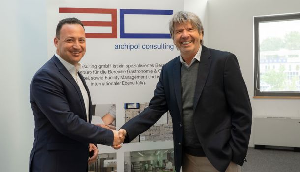 Die beiden geschäftsführenden Gesellschafter schütteln sich die Hand vor einem Aufsteller von Archipol Consulting.