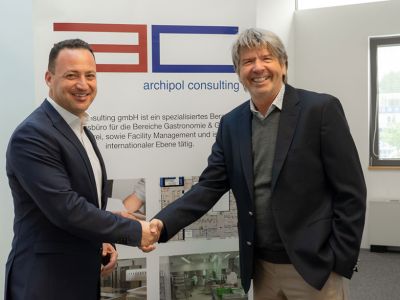 Die beiden geschäftsführenden Gesellschafter schütteln sich die Hand vor einem Aufsteller von Archipol Consulting.