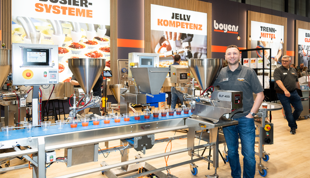 Ein Mann steht hinter einer Portioniermaschine, die neben einem Fließband montiert wurde, um Lebensmittelzutaten in Bechern auf dem Fließband zu portionieren.